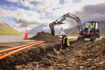 Construction scene in a mountainous area, showing a worker in a hard hat and reflective vest inspecting or working in a trench beside an excavator. Bright orange conduit pipes lie in the trench, while the excavator, operated by another worker in a similar outfit, is actively digging or moving dirt. Traffic cones are positioned near the trench for safety, and clouds partially cover the sky, casting soft light over the scenic background of rolling green hills and distant mountains.