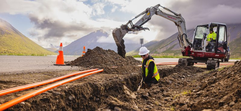 Construction scene in a mountainous area, showing a worker in a hard hat and reflective vest inspecting or working in a trench beside an excavator. Bright orange conduit pipes lie in the trench, while the excavator, operated by another worker in a similar outfit, is actively digging or moving dirt. Traffic cones are positioned near the trench for safety, and clouds partially cover the sky, casting soft light over the scenic background of rolling green hills and distant mountains.