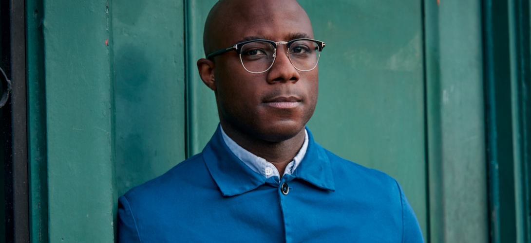  Filmmaker Barry Jenkins, a bald Black man wearing glasses and a vibrant blue button-up jacket over a white collared shirt, stands against a deep green door. 