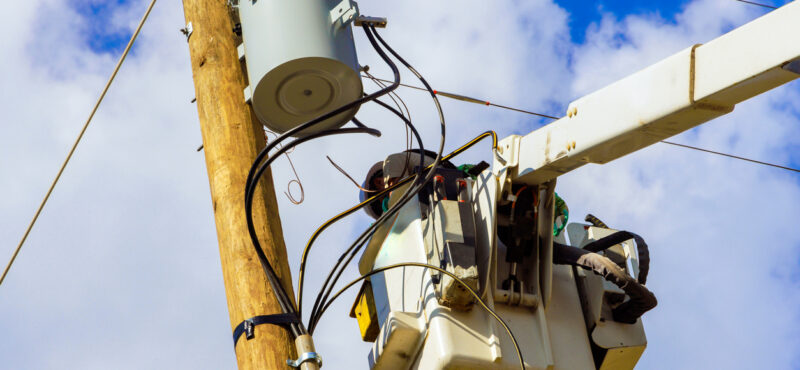 Telecom worker in a bucket truck repairing or installing lines on a utility pole with a transformer, under a partly cloudy sky.