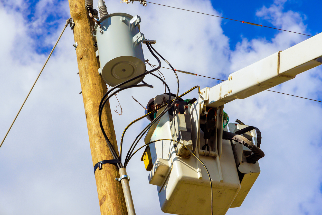 Telecom worker in a bucket truck repairing or installing lines on a utility pole with a transformer, under a partly cloudy sky.